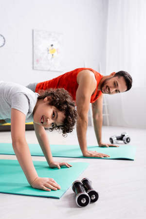 Smiling arabian kid working out on fitness mat near father and dumbbells on blurred background