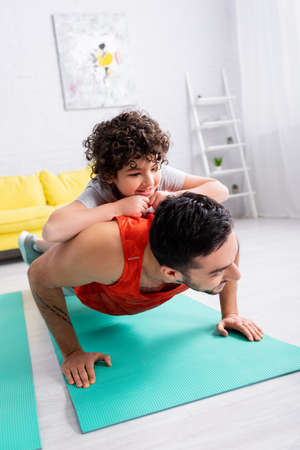 Smiling arabian kid on back of father training on fitness mat at home