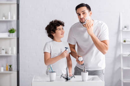 Smiling arabian boy holding razor near father with shaving foam in bathroomの写真素材
