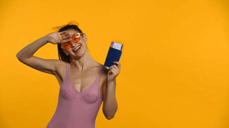 Smiling woman in swimsuit and sunglasses holding passport isolated on yellow