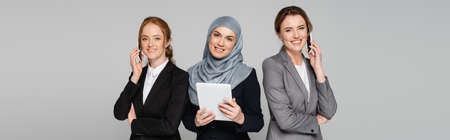 Smiling multiethnic businesswomen talking on smartphones and using digital tablet isolated on gray, bannerの写真素材