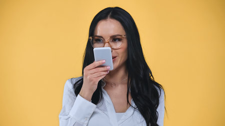 brunette businesswoman in eyeglasses looking at smartphone isolated on yellow