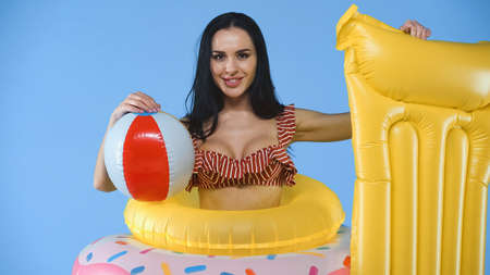 happy woman with swim rings, inflatable ball and mattress smiling at camera isolated on blue