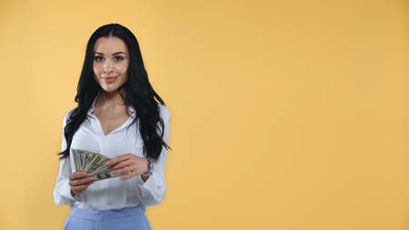 successful businesswoman holding dollars while smiling at camera isolated on yellowの写真素材