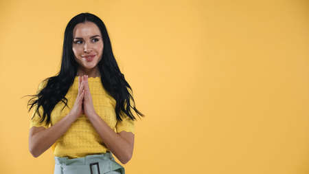 positive brunette woman standing with praying hands isolated on yellowの写真素材