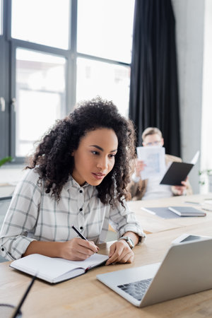African american businesswoman writing on notebook near laptop on blurred foreground in office