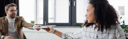 Smiling african american businesswoman giving documents to colleague on blurred background, bannerの写真素材