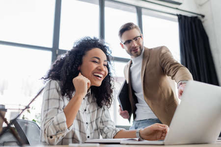 Smiling african american businesswoman looking at laptop near colleague on blurred backgroundの写真素材