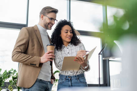 Smiling african american businesswoman holding pen and paper folder near businessman with takeaway drinkの写真素材