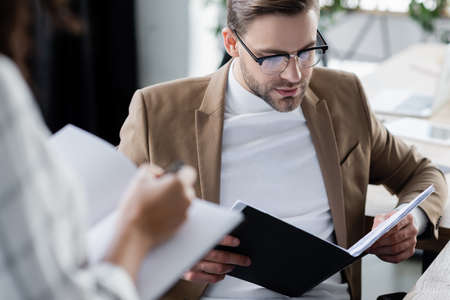 Businessman looking at documents near colleague with notebook on blurred foreground