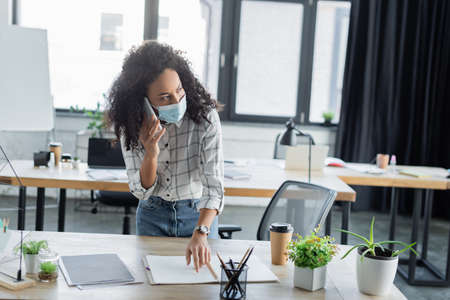 african american businesswoman in medical mask pointing at notebook while talking on mobile phoneの写真素材