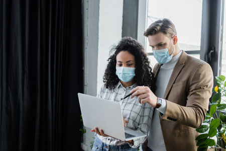 young businessman in protective mask pointing at laptop in hands of african american coworkerの写真素材