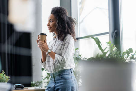 Positive african american businesswoman holding takeaway drink near table in officeの写真素材