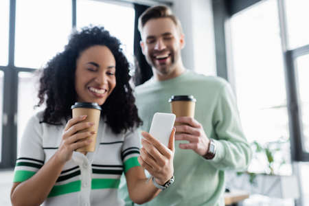 cheerful interracial business colleagues looking at smartphone while holding coffee to go, blurred backgroundの写真素材