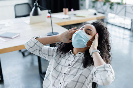 exhausted african american businesswoman in medical mask touching head while sitting with closed eyesの写真素材