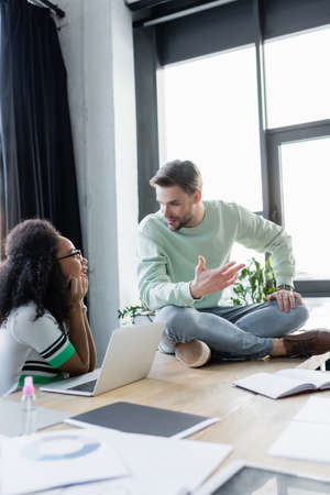 Interracial business people talking near papers and laptop in office