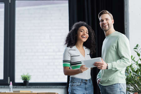 happy interracial business colleagues looking at camera while holding digital tablet
