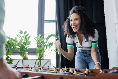 excited african american businesswoman showing win gesture near table football, blurred foreground