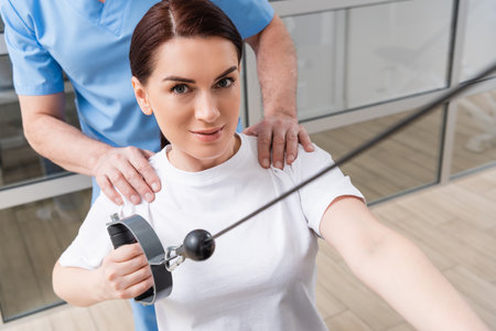 smiling woman looking at camera while working out in rehabilitation center near trainer