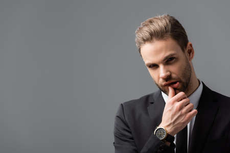 pensive businessman looking at camera while touching lip isolated on grayの写真素材