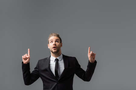 astonished businessman looking up and pointing with fingers isolated on grayの写真素材