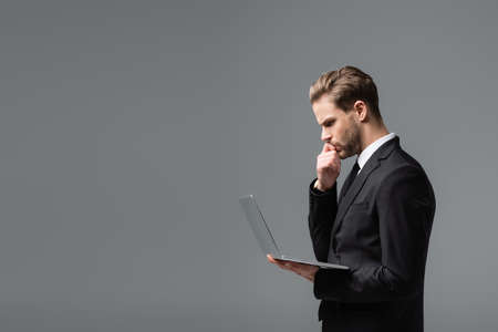 side view of thoughtful businessman looking at laptop isolated on grayの写真素材