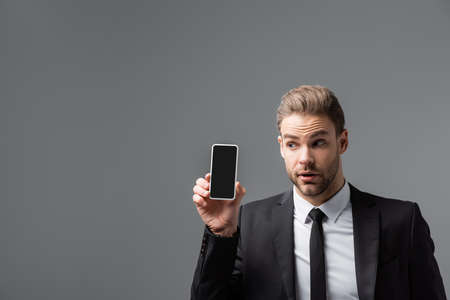 curious businessman holding mobile phone with blank screen isolated on grayの写真素材