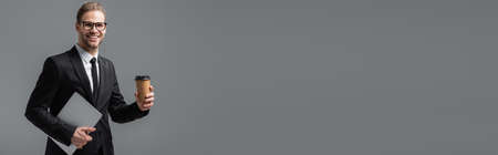 businessman in eyeglasses holding coffee and laptop isolated on gray, bannerの写真素材