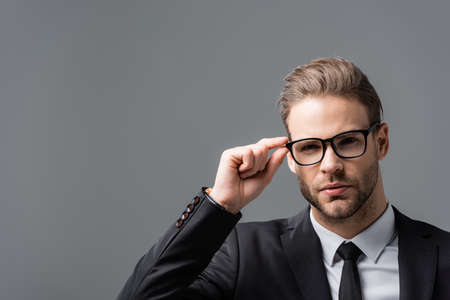 young businessman touching eyeglasses while looking at camera isolated on grayの写真素材