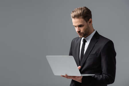 young, concentrated businessman using laptop isolated on grayの写真素材