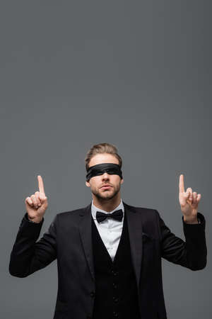 young, blindfolded businessman pointing up with fingers isolated on grayの写真素材