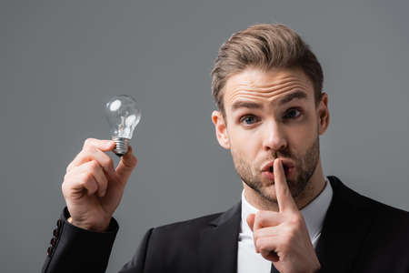 thrilled businessman holding light bulb while showing secret gesture isolated on grayの写真素材