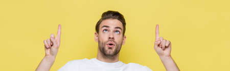 surprised man pointing with fingers while looking up isolated on yellow, bannerの写真素材