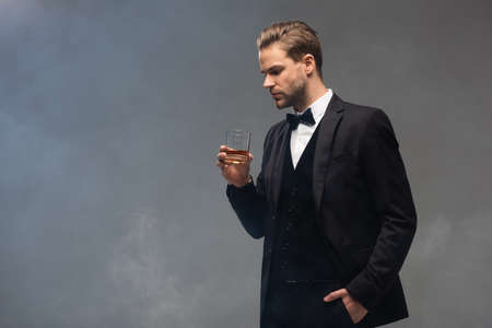 young businessman holding whiskey while standing with hand in pocket on gray background with smoke