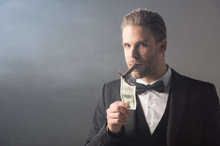 businessman lighting cigar from dollar banknote while looking at camera on gray background with smokeの写真素材