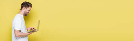 side view of smiling man typing on laptop on yellow, bannerの写真素材