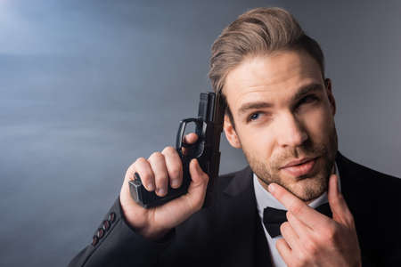 thoughtful businessman touching face while holding gun near head on gray background with smokeの写真素材