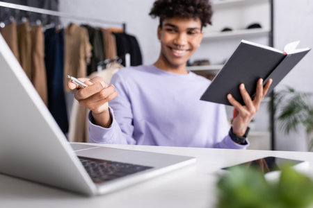 Pen in hand of african american businessman with notebook on blurred foreground near gadgetsの写真素材