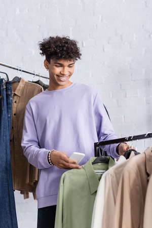 Smiling african american owner of showroom holding smartphone near shirts on hangersの写真素材
