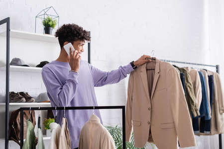 Positive african american owner of showroom talking on smartphone and looking at jacket on hangerの写真素材