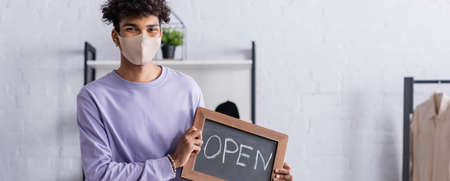 African american small business owner in protective mask holding chalkboard with open lettering,...