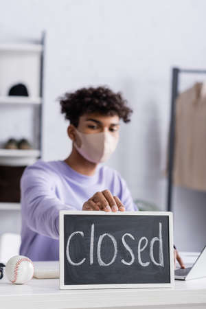 Chalkboard with closed lettering near african american owner of showroom in protective mask working on blurred backgroundの写真素材