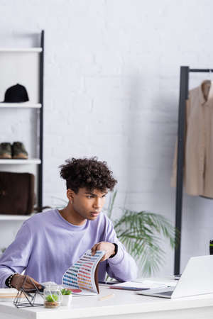 Young african american owner of showroom holding colorful swatches and looking at laptop
