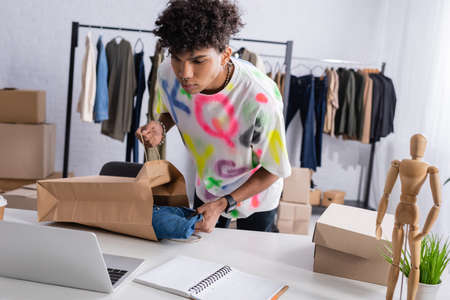 African american owner of showroom holding shopping bag and shirt near laptop and box