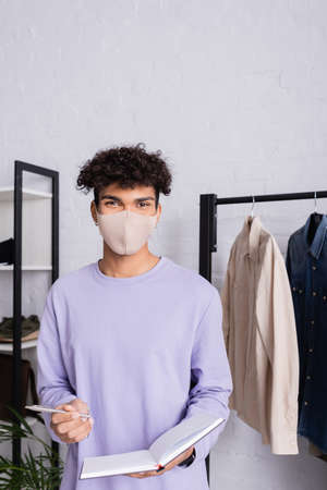 African american showroom proprietor in protective mask holding pen and notebook near clothesの写真素材