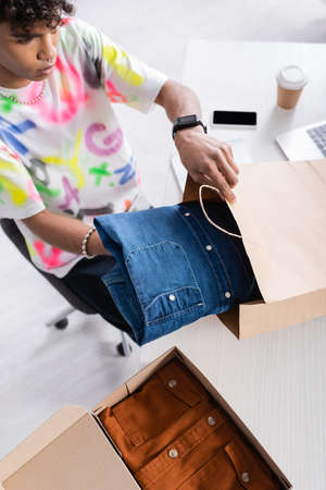 High angle view of african american owner of showroom putting shirt in shopping bag
