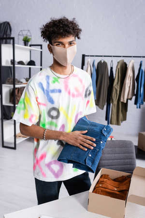 African american owner of showroom in protective mask holding shirt near carton box