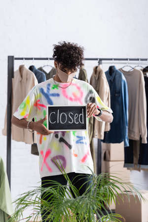 Stylish african american owner of showroom in protective mask holding chalkboard with closed...