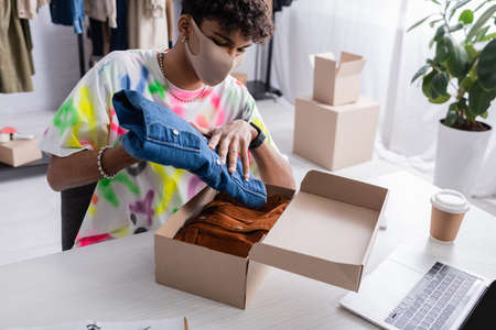 African american showroom owner in protective mask putting clothes in carton box near laptop and...