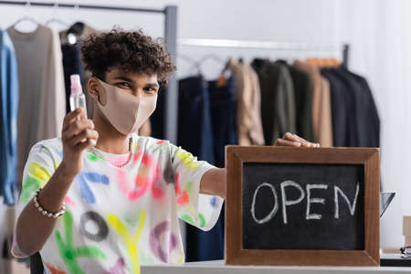 African american owner of showroom in protective mask holding hand sanitizer and chalkboard with...